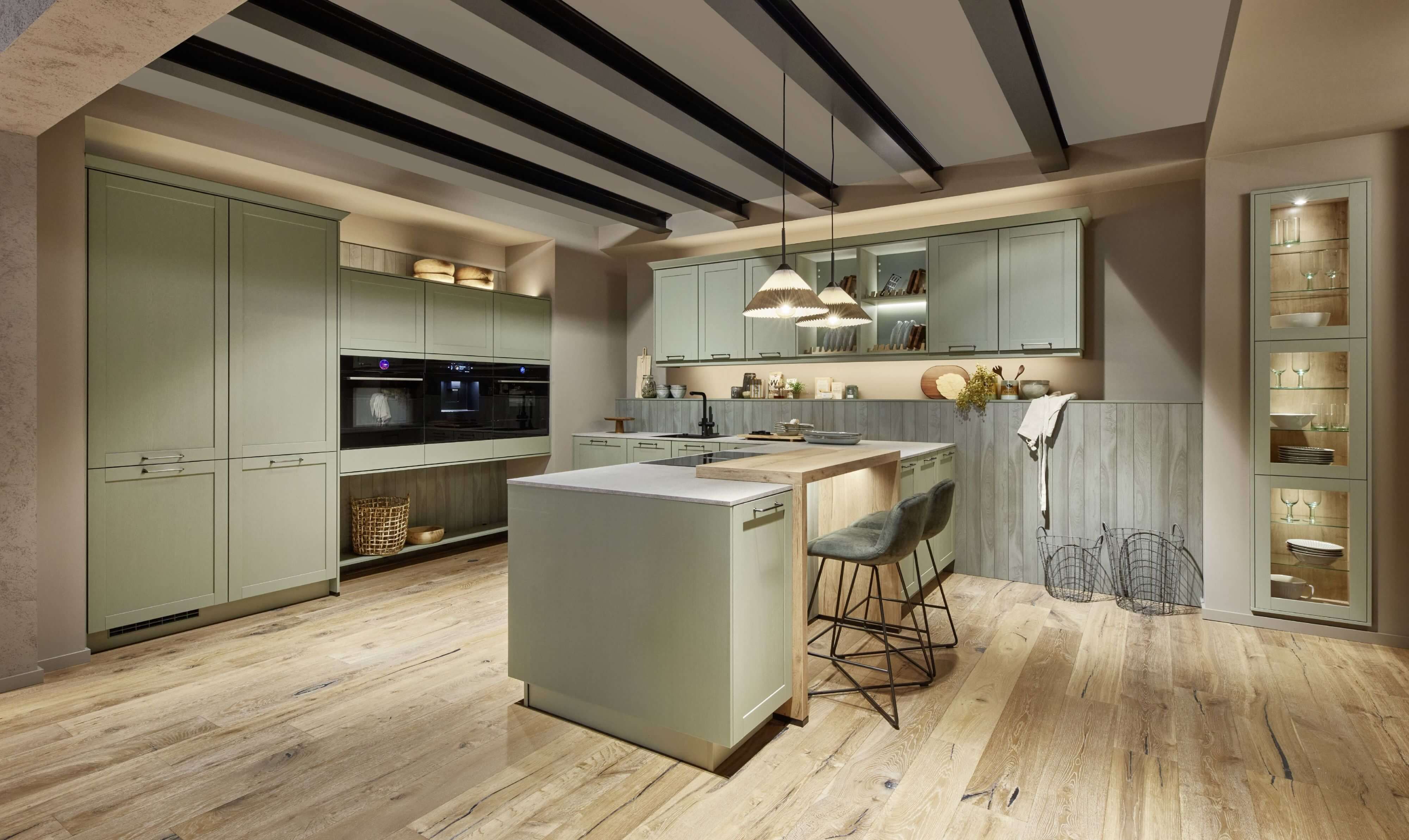 Hochwertige Landhausstlküche mit klassischen Rahmenfronten in saftem Grün. Der natürlich Holzboden, die offenen Regale und die indirete Beleuchtung unterstreichen die wame wohnliche Atmosphäre.