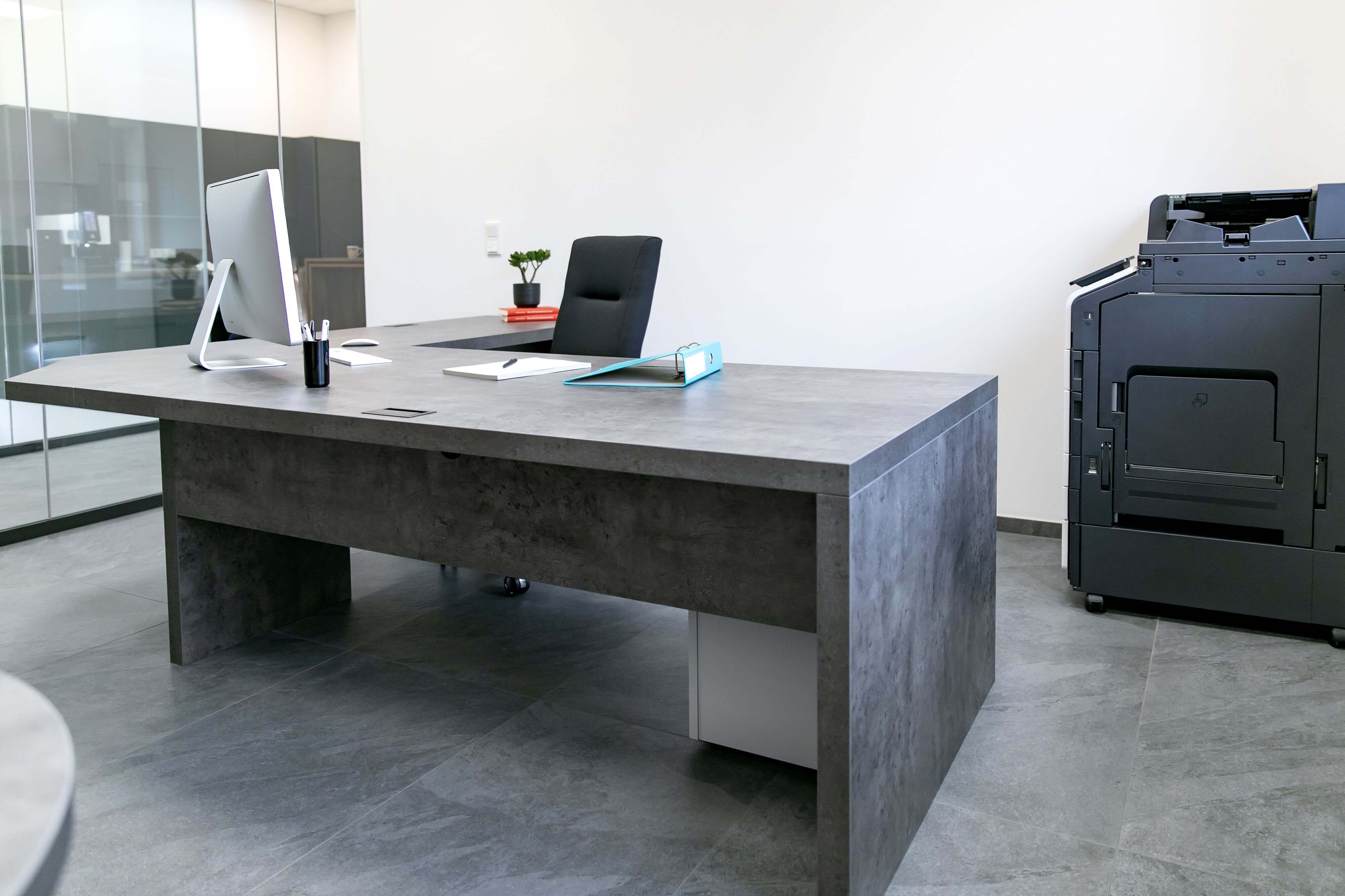 Moderner Büroarbeitsplatz mit großem grauen Schreibtisch in Betonoptik, Computer, blauem Aktenordner und Multifunktionsdrucker im hellen Raum