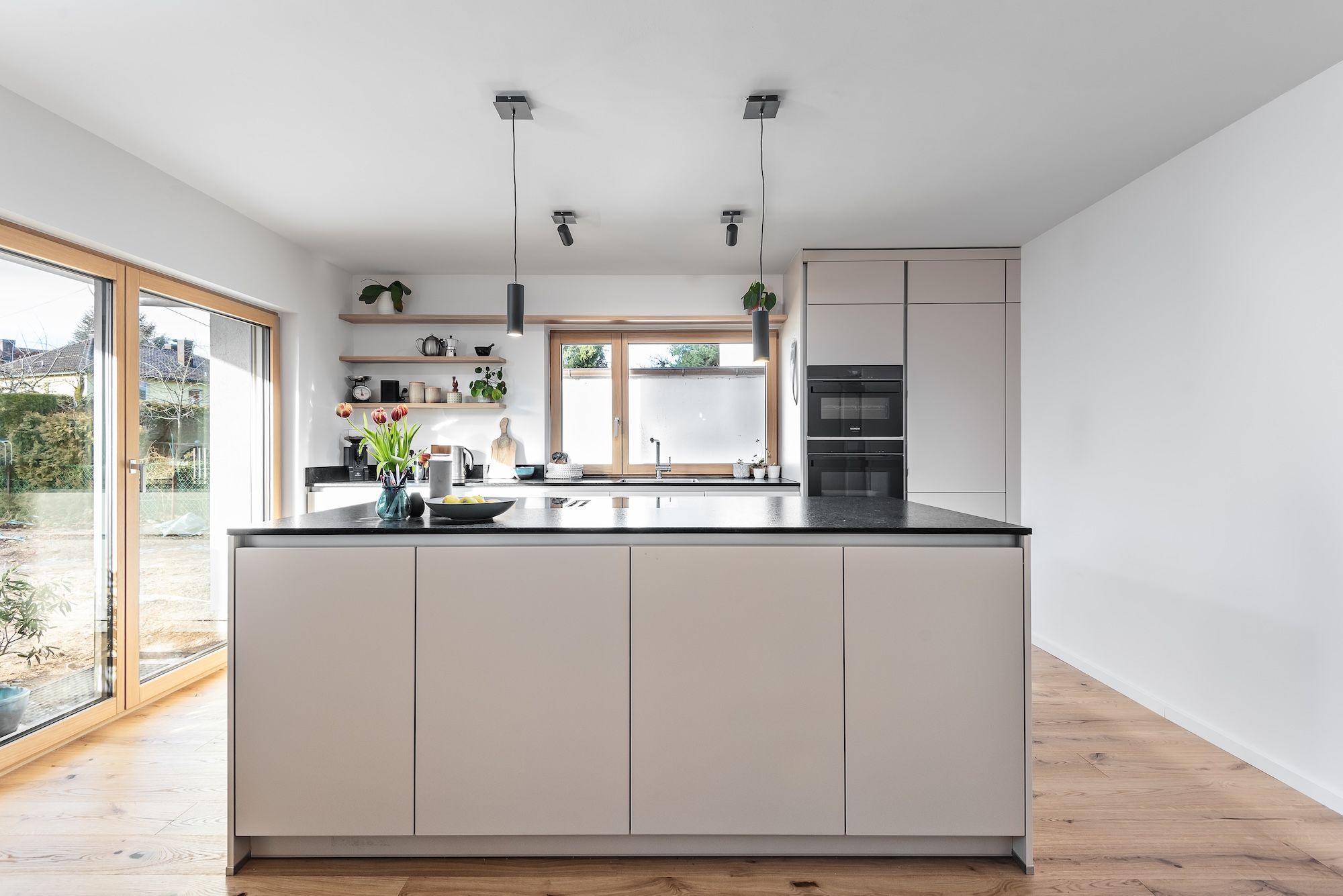 Moderne Wohnküche mit Küchenisnel, klarer, reduzierter Gestaltung. Helle grifflose Fronten in warmen Naturtönen werden durch die dunkle Granitarbeitsplatte stilvoll kontrastiert.