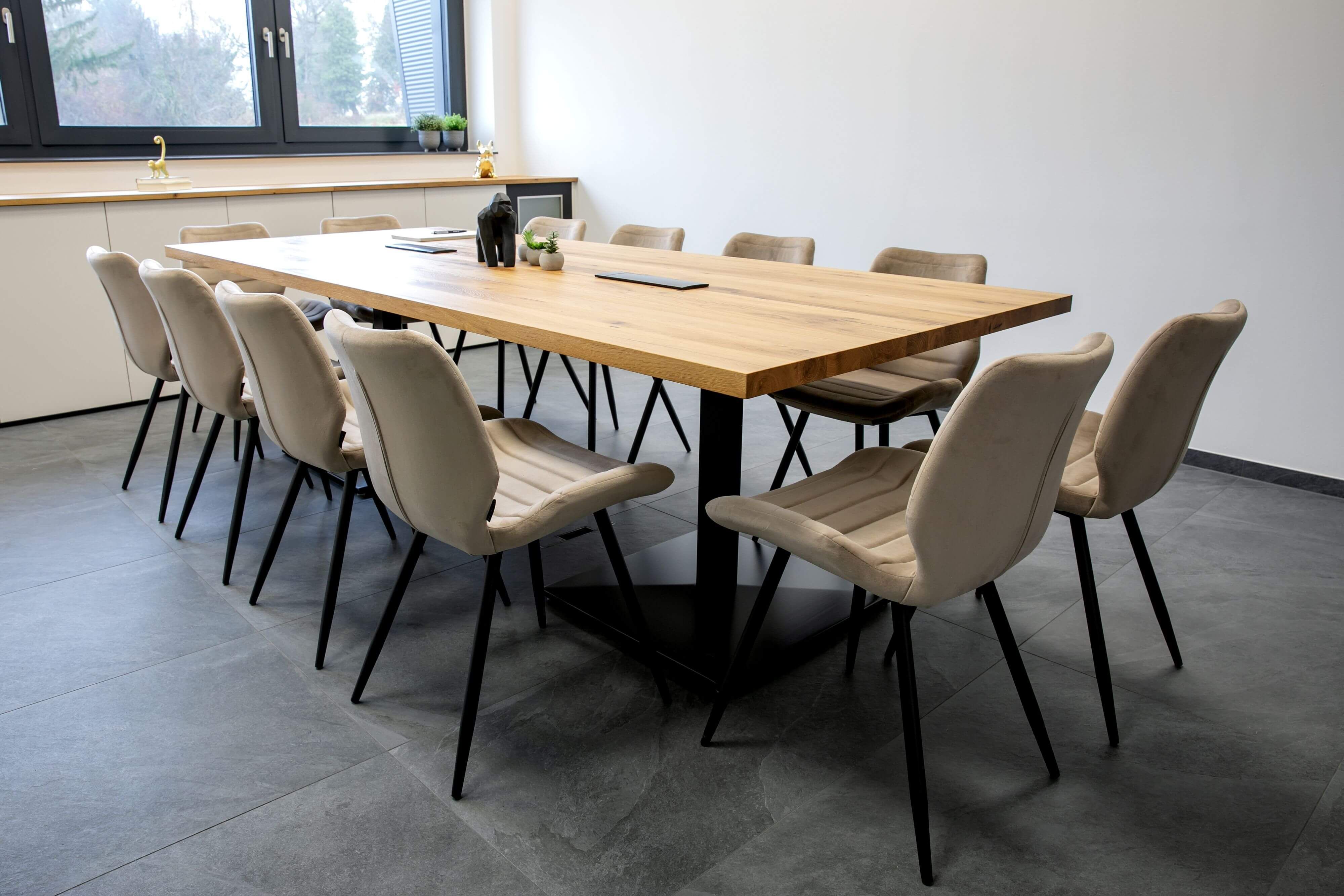 Großer Massivholztisch aus Eiche mit schwarzem Metallgestell und beigen Polsterstühlen in einem hellen Konferenzraum oder Esszimmer mit grauen Fliesen