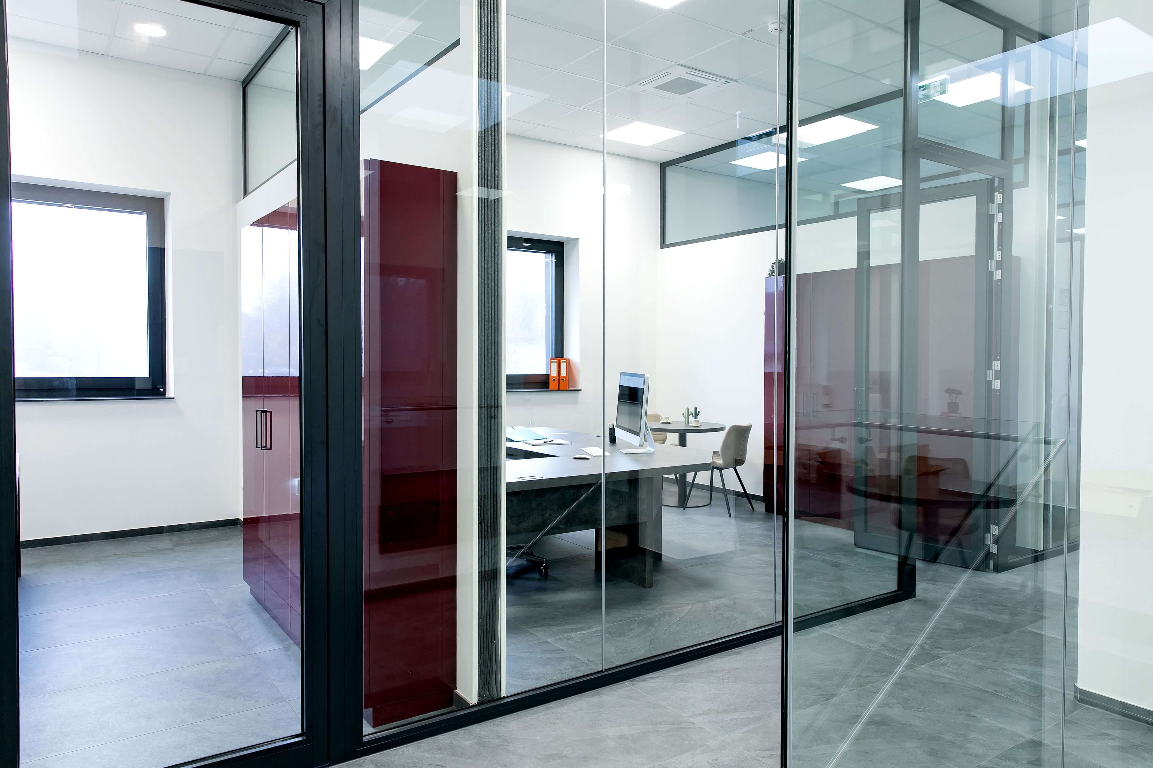 Blick durch transparente Glastrennwände in ein modernes Büro mit grauem Schreibtisch, Computer, weinrotem Einbauschrank und grauen Bodenfliesen