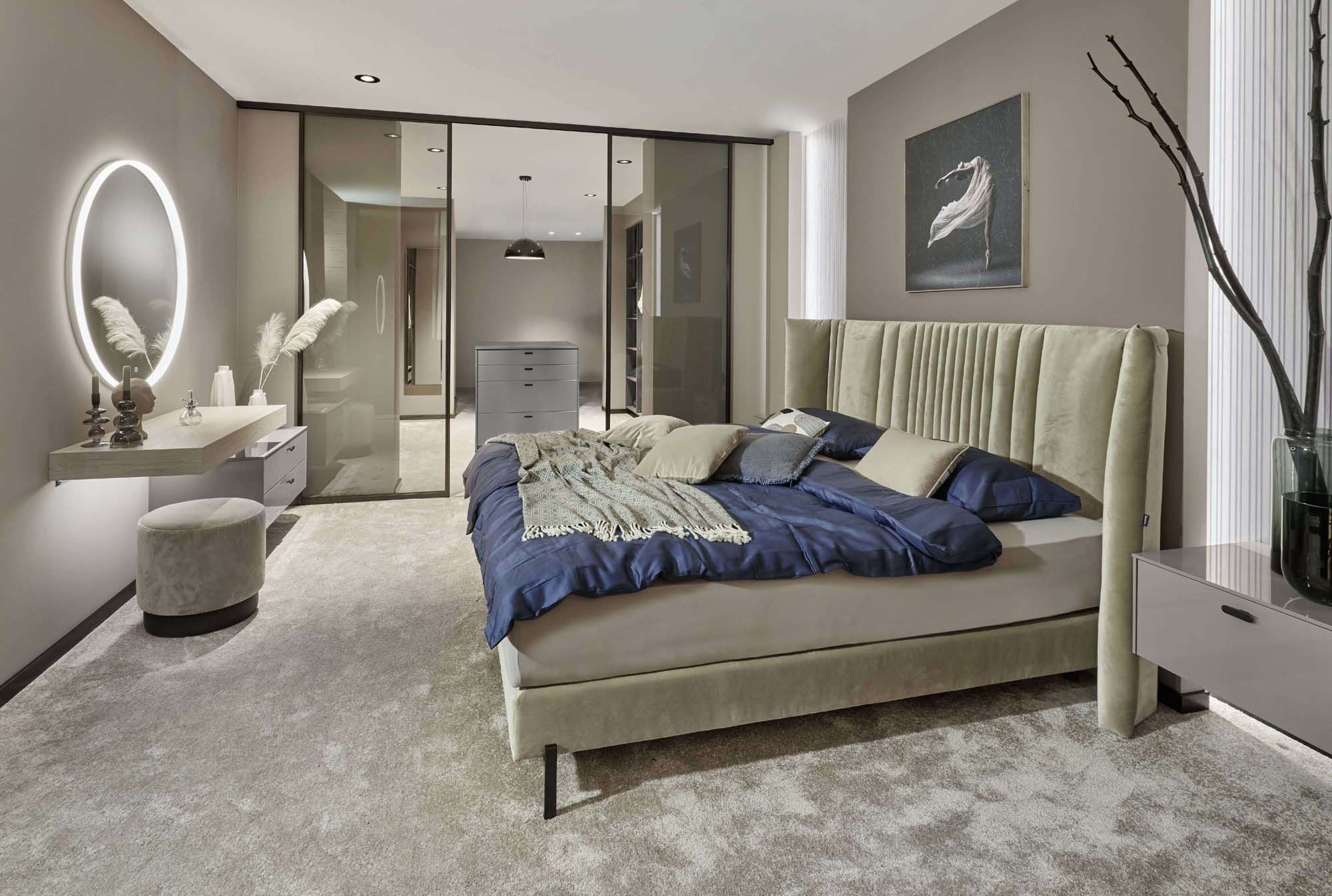 Elegantes Schlafzimmer mit gepolstertem Bett, ruhiger Farbwelt und integrierter Ankleide hinter Glasfronten. Die durchdachte Raumplanung verbindet Schlaf- und Stauraumbereich harmonisch und schafft eine luxuriöse, entspannte Wohnatmosphäre.